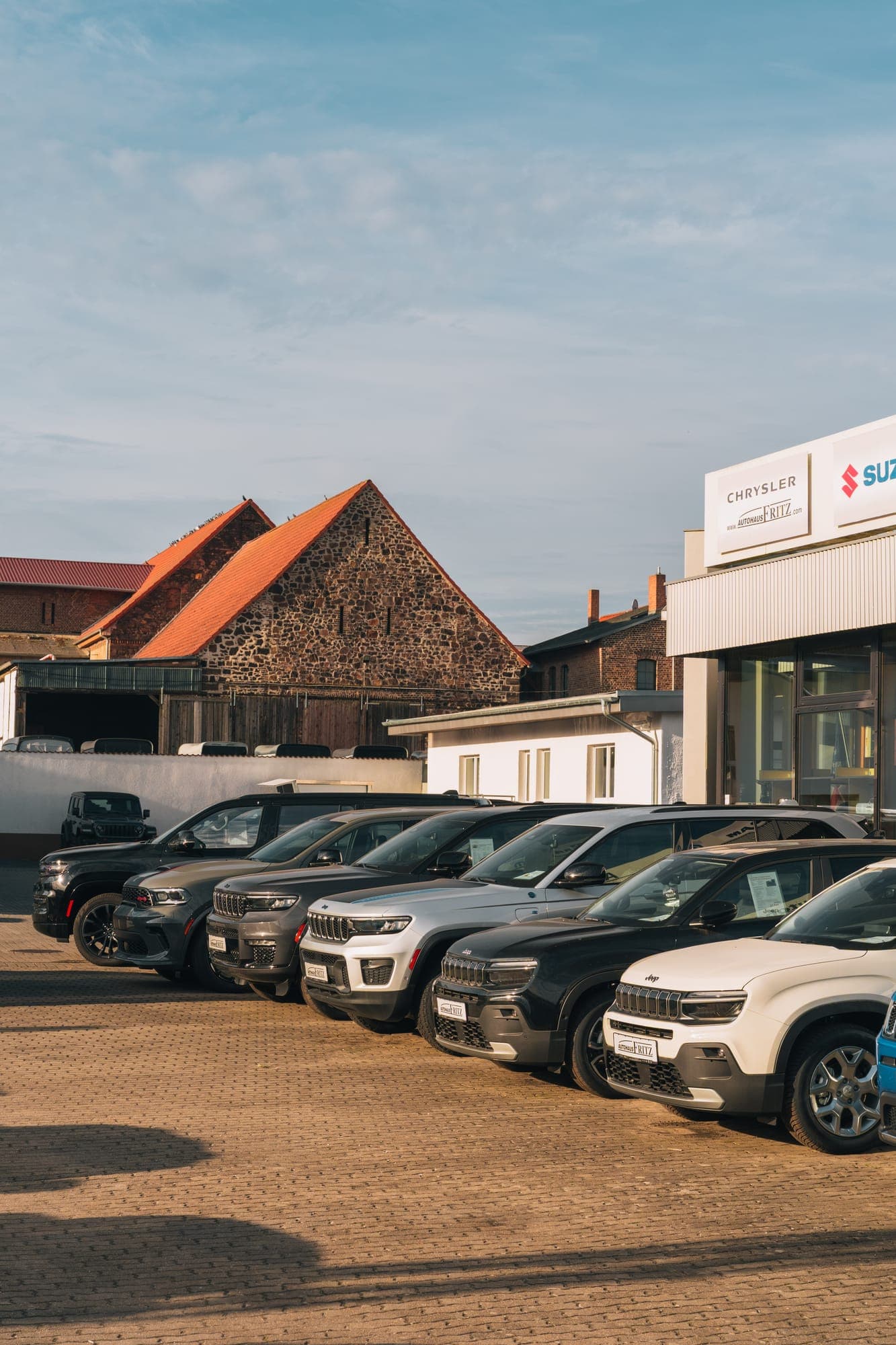 The dealership yard in Oppin: Jeep and Dodge vehicles in a row in front of the sales building with brand signs, an old fieldstone barn behind them